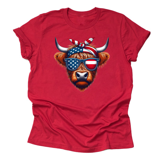 A red short-sleeved unisex T-shirt featuring a graphic of a Highland cow with American flag patterned sunglasses and a bow.